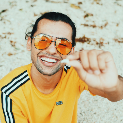 A man in a yellow shirt wearing orange aviator sunglasses while holding a shark tooth on the beach.