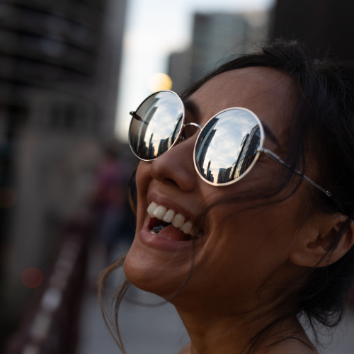 A woman on a city side walk is wearing a round pair of reflective sunglasses while looking towards the sky.